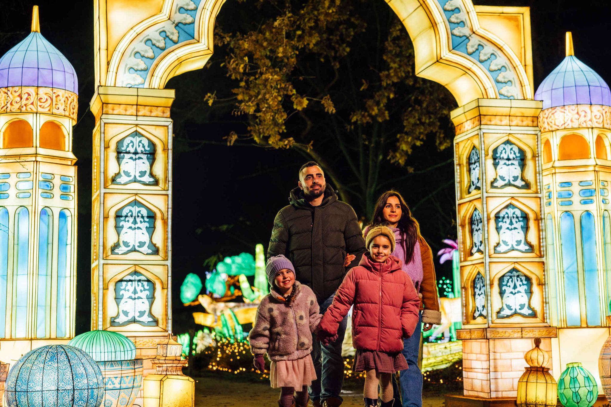A family of four, dressed in winter clothes, stands under a brightly lit, decorative archway at night with colorful illuminated structures around them.