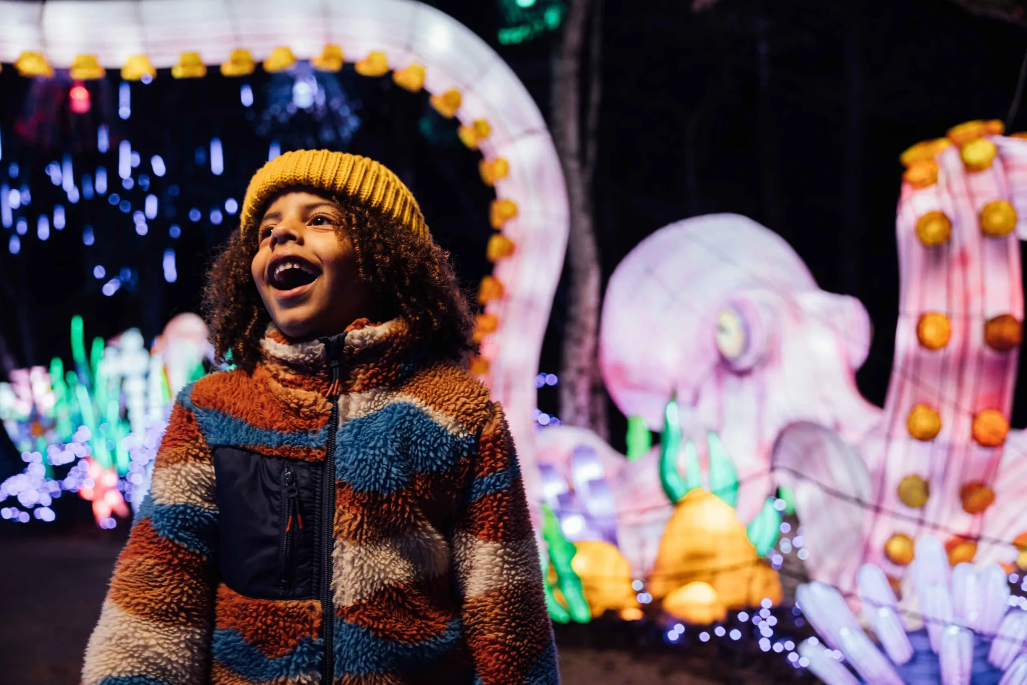 A child wearing a colorful jacket and yellow hat smiles joyfully at a brightly lit outdoor display featuring large, illuminated sea creatures and underwater decorations at night.