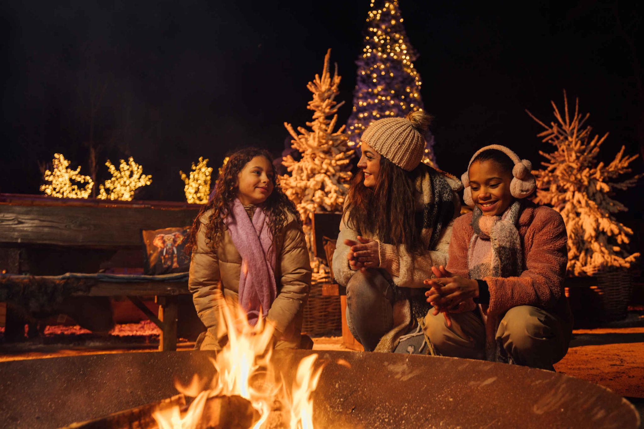 Three people dressed in winter clothing sit around a fire pit at night, smiling and talking. Behind them are lit Christmas trees and festive lights, creating a warm, holiday atmosphere.