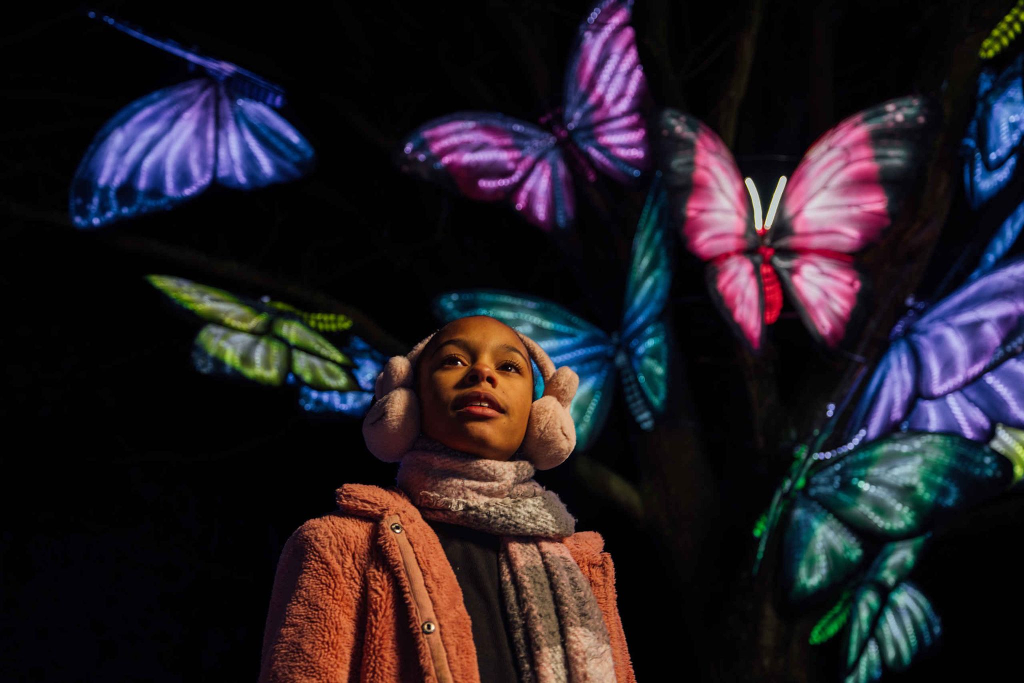 A child wearing earmuffs and a scarf looks up, standing in front of a tree decorated with large, brightly lit, colorful butterfly lanterns at night.