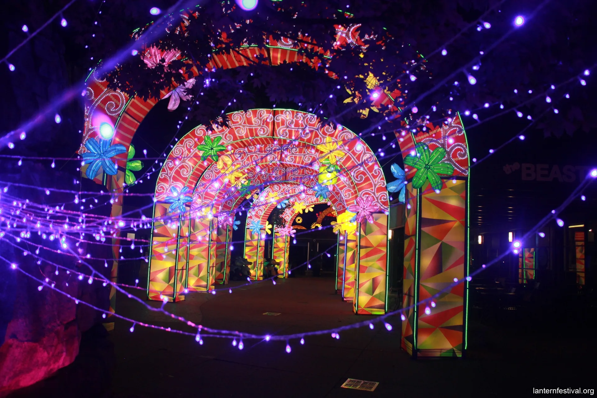 An illuminated outdoor tunnel made of colorful archways and pillars, featuring geometric patterns and floral light sculptures. Purple string lights hang on both sides.