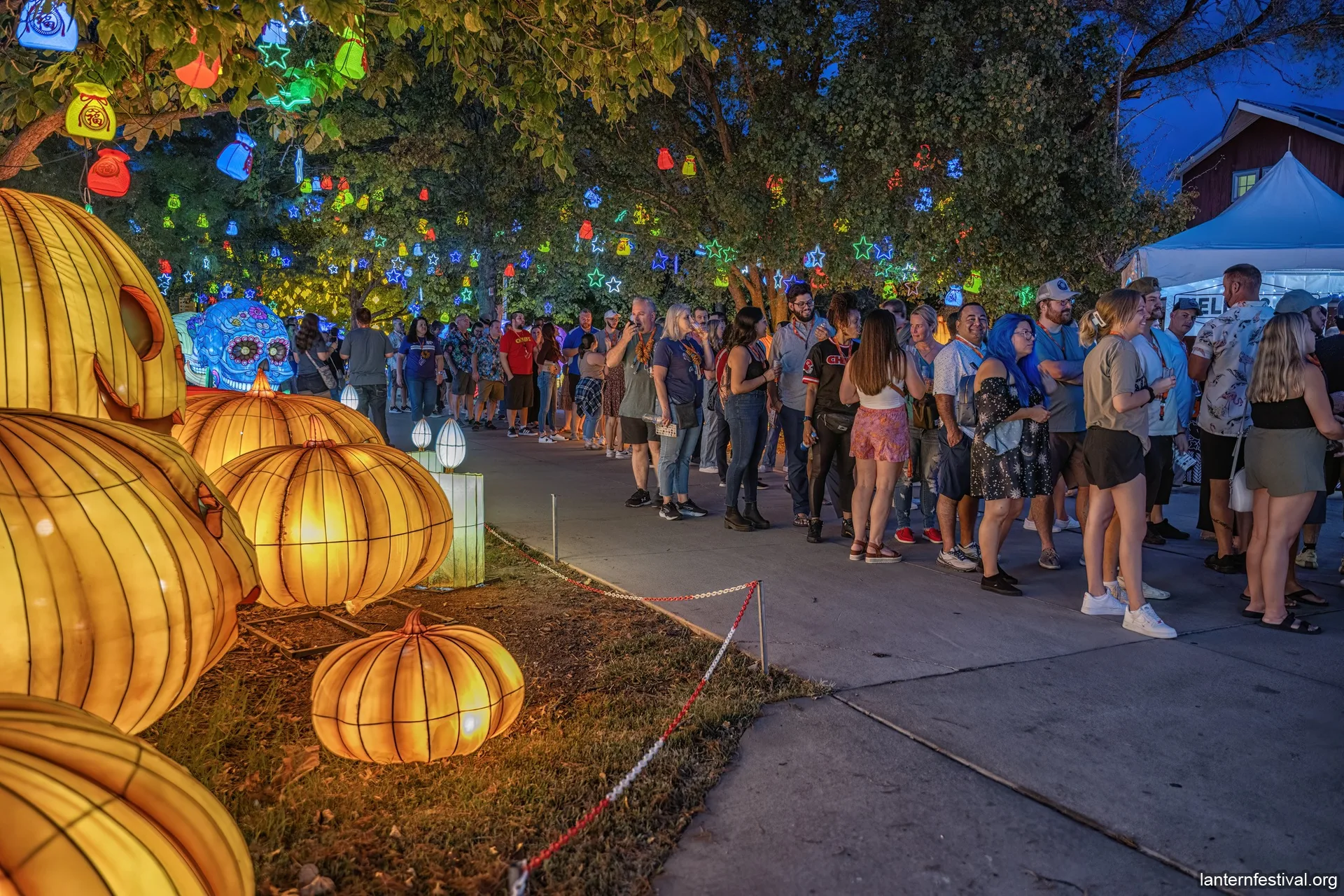 An outdoor lantern festival at dusk, featuring large pumpkin and skull lanterns. Many colorful lanterns hang from trees above a crowd.